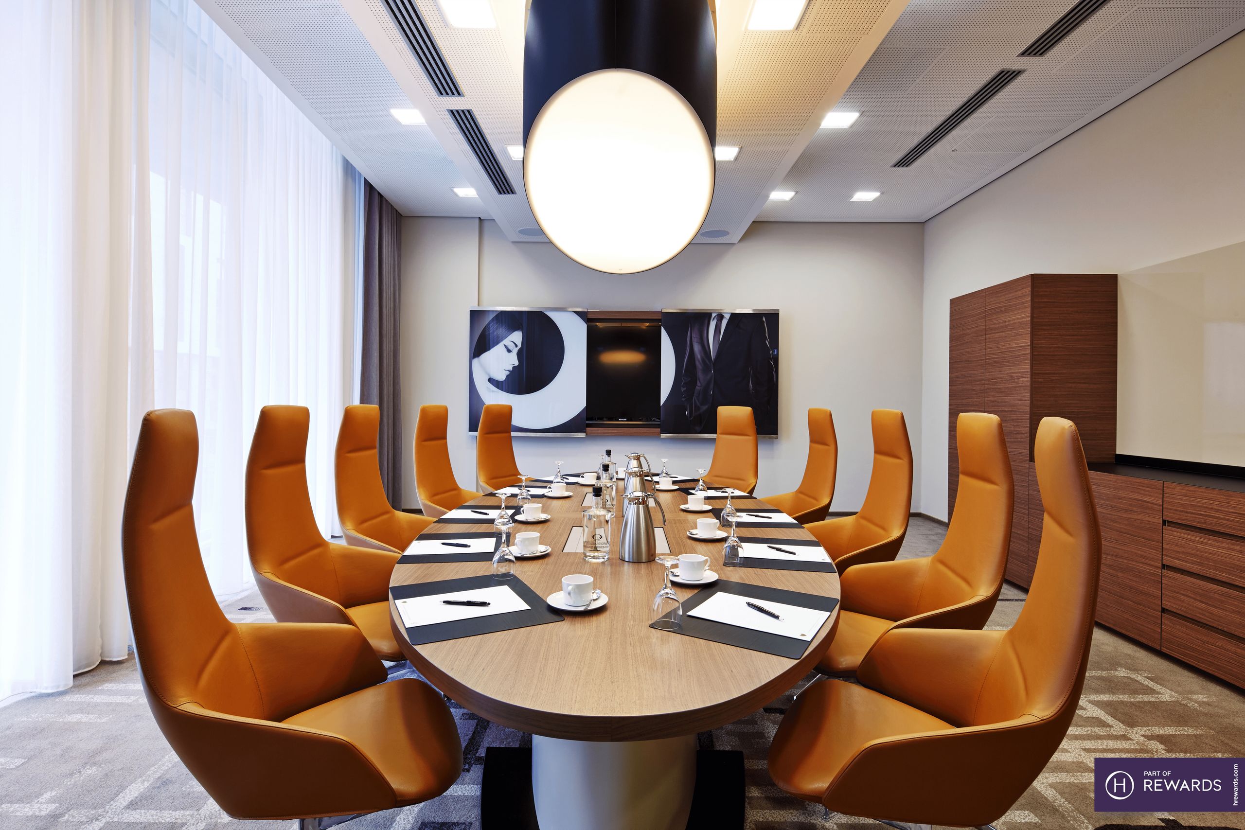 A modern conference room with an oval table surrounded by orange chairs and a large overhead light.