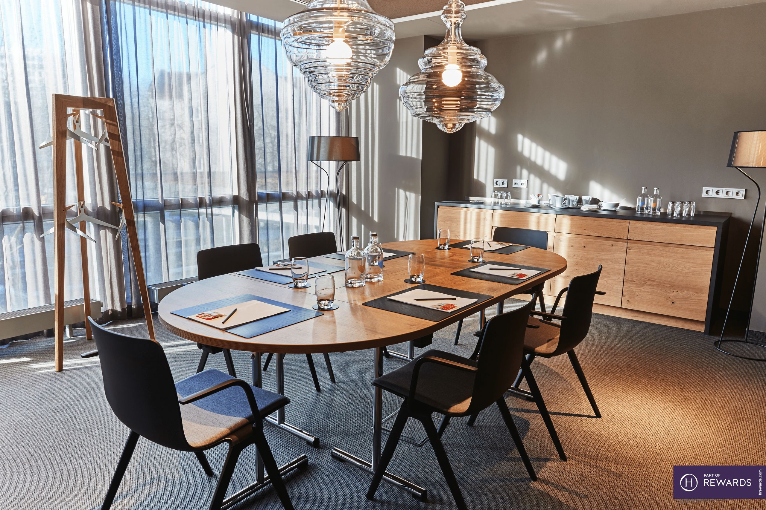 A modern conference room with a large wooden table, black chairs, pendant lights, and natural…