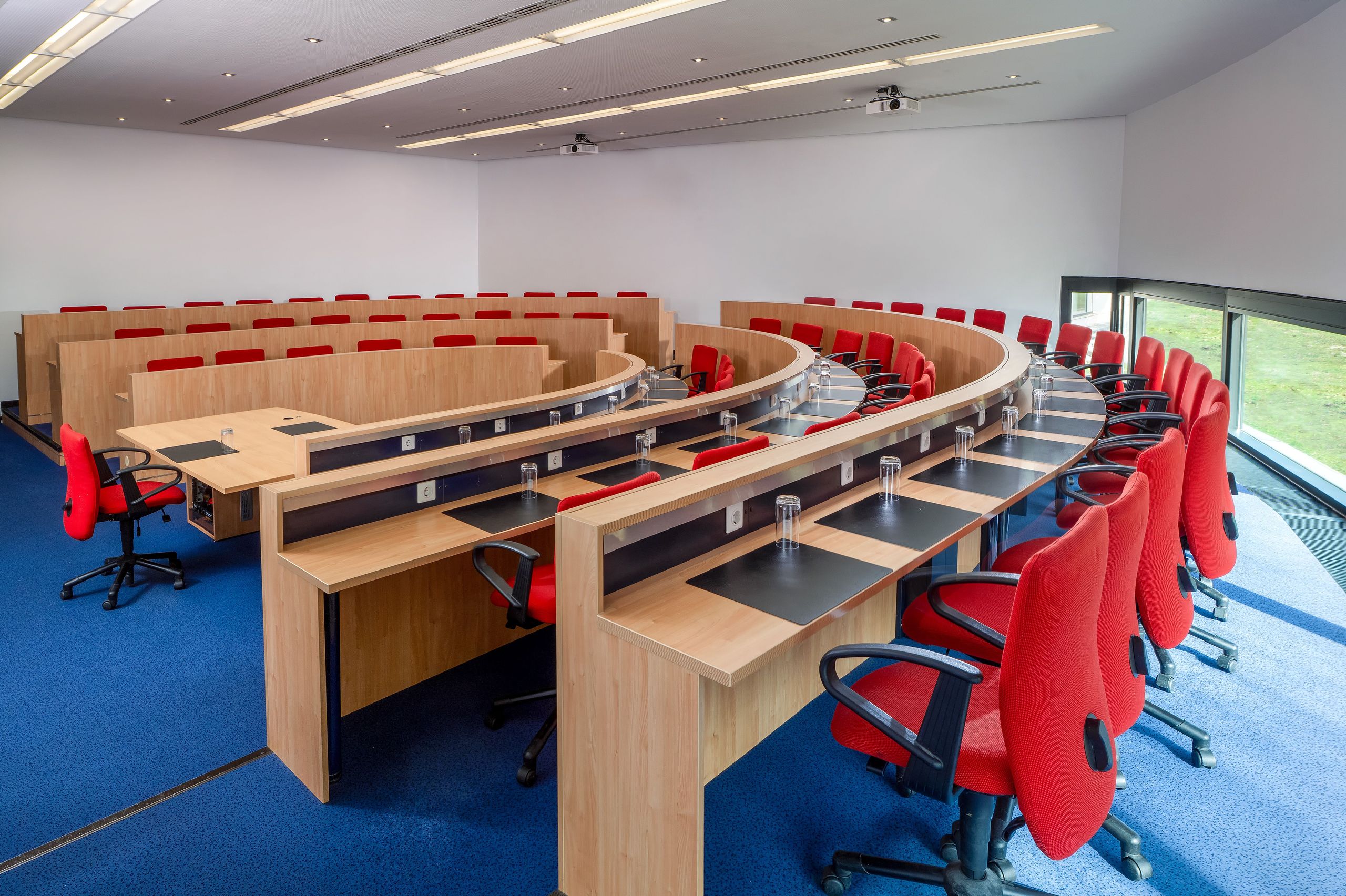 Ein moderner Hörsaal oder Konferenzraum mit gebogenen Reihen aus Holzpulten und roten Drehstühlen.