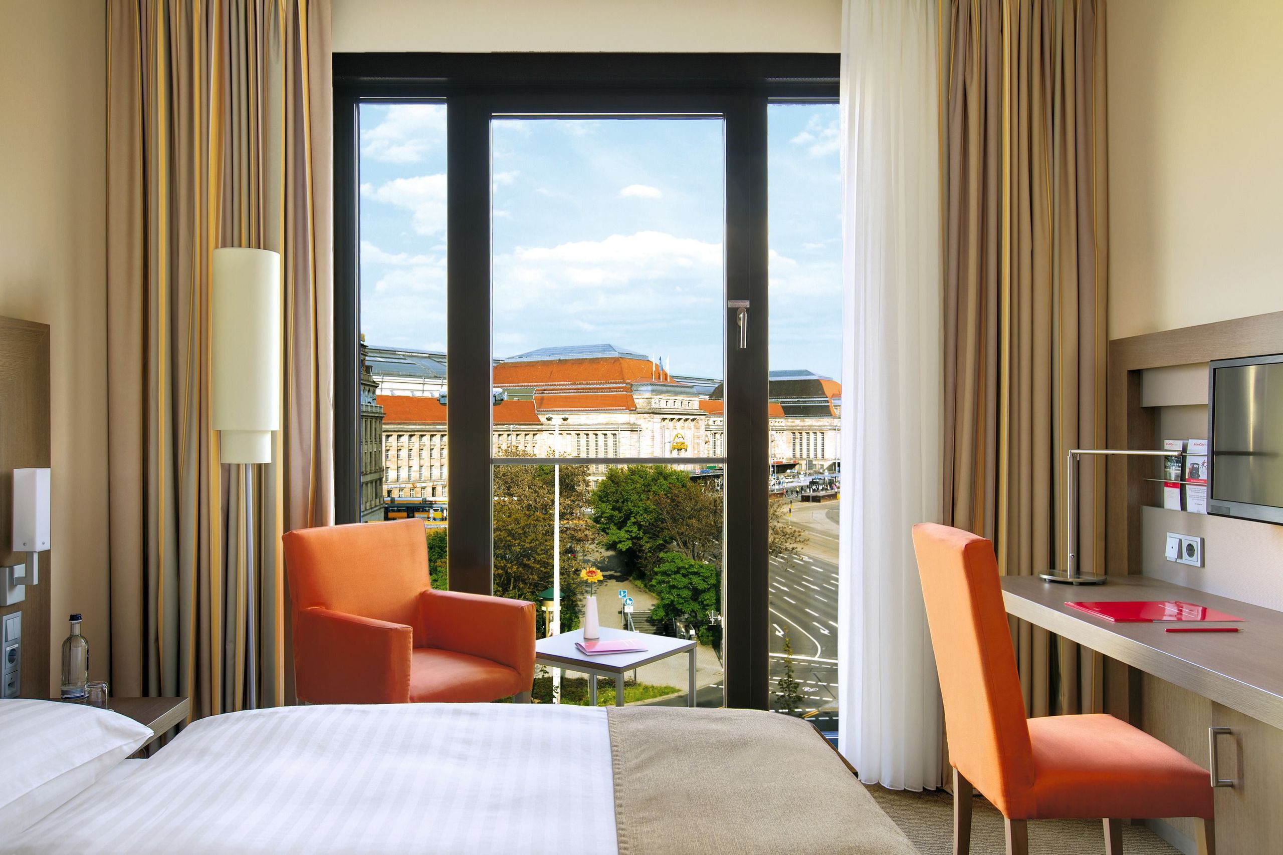 Interior de una habitación de hotel con una cama, dos sillas naranjas, un escritorio y una ventana…