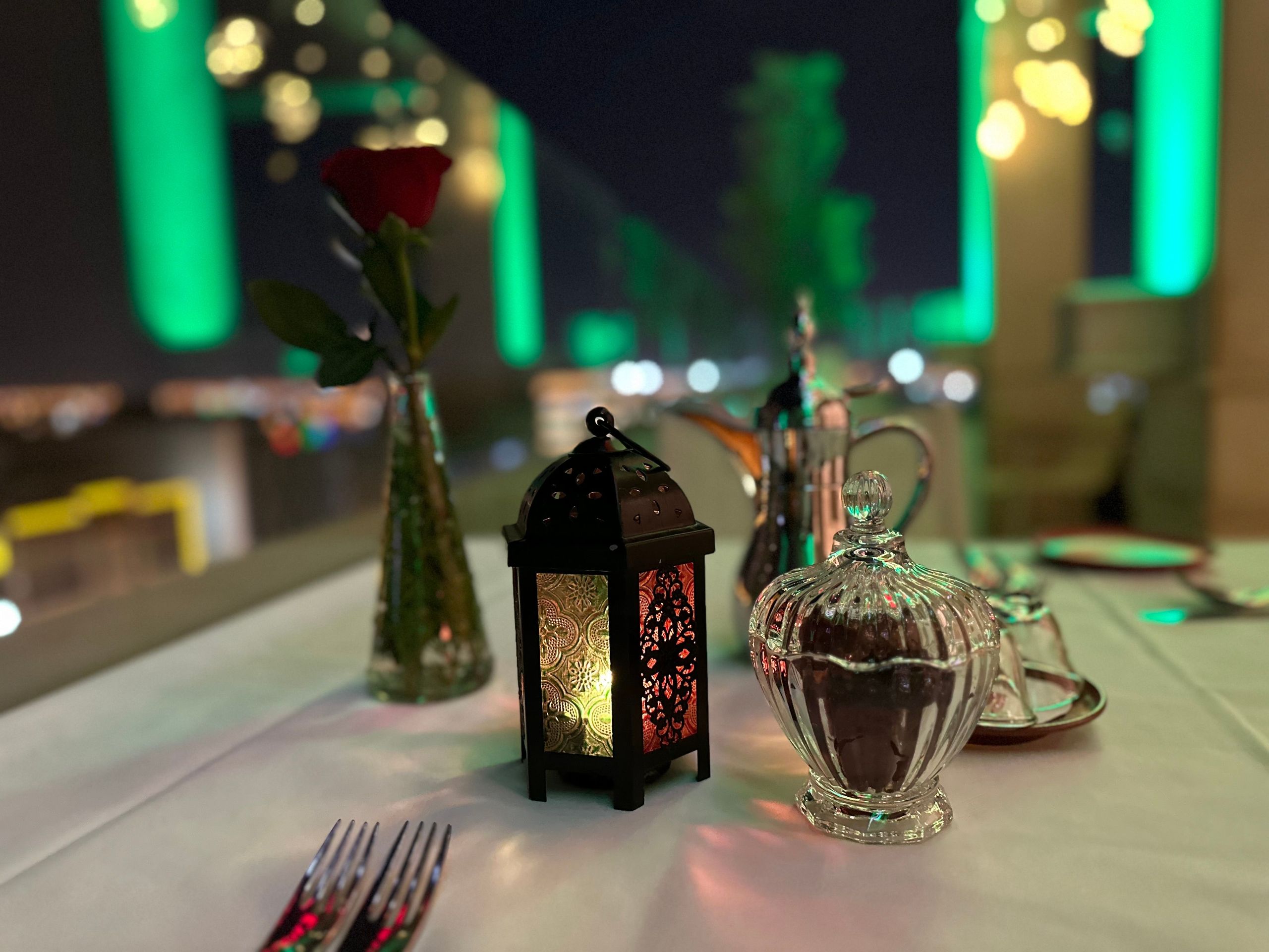 Una mesa con poca luz, con una linterna decorativa, un jarrón de vidrio con una rosa roja y…