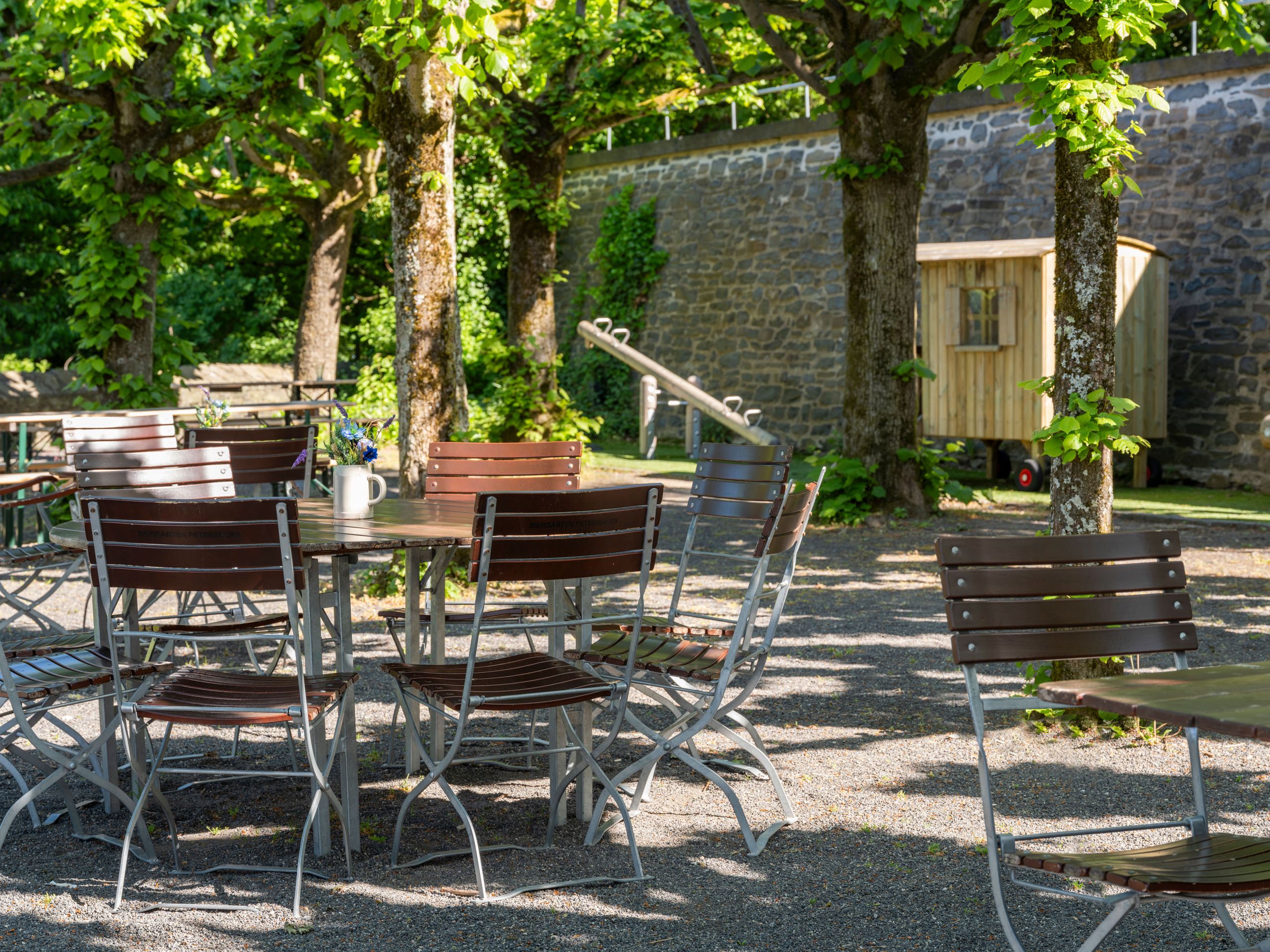 An outdoor seating area with metal and wooden chairs and tables under trees.