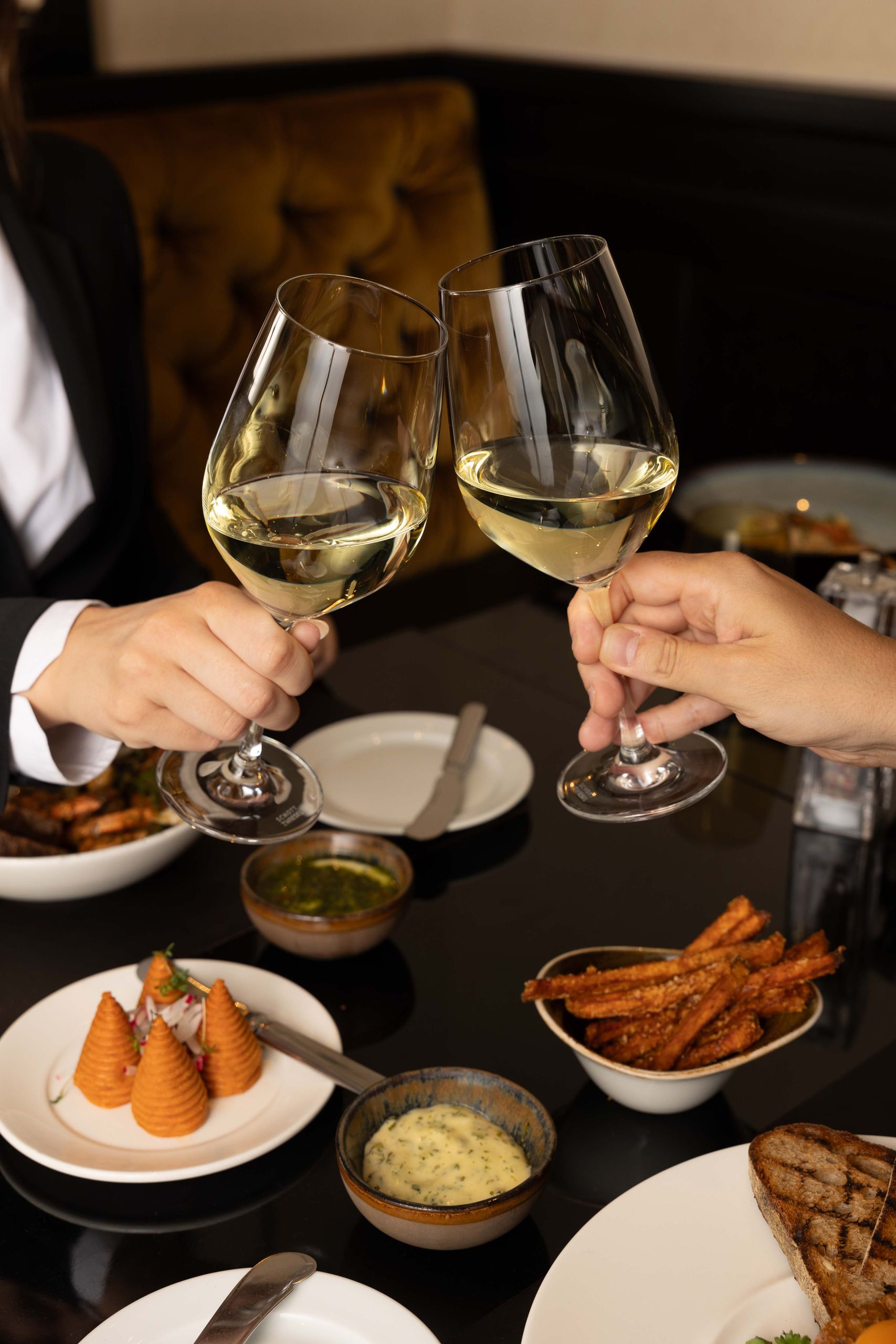 Två personer skålar med glas vitt vin över ett bord med olika rätter.