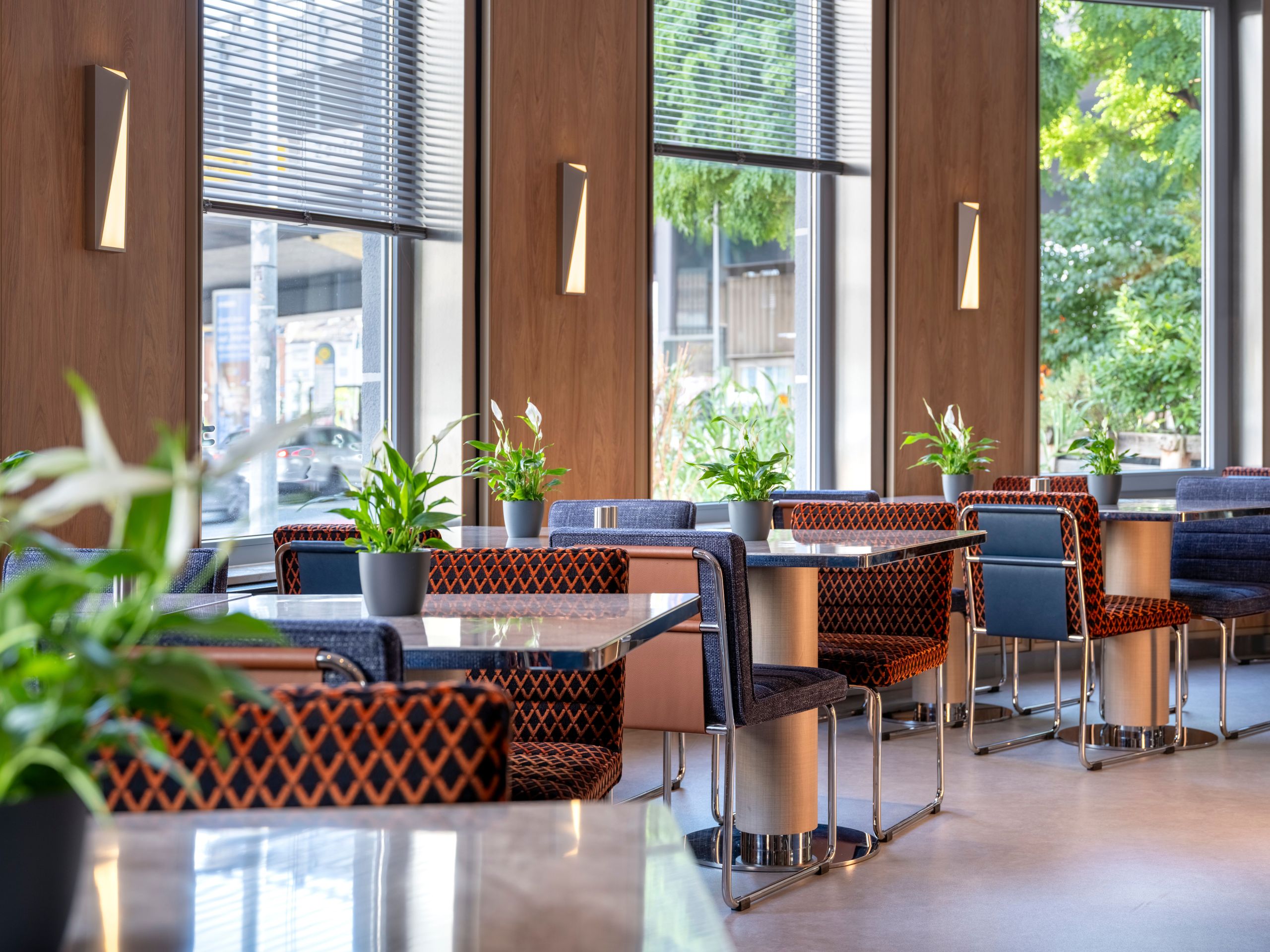 The photo shows a modern indoor seating area with tables, chairs, and potted plants near large…