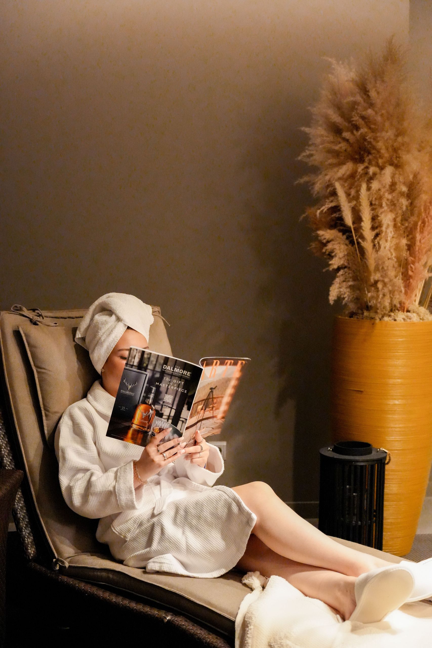 A person in a bathrobe and towel turban is sitting on a lounge chair and reading a magazine.