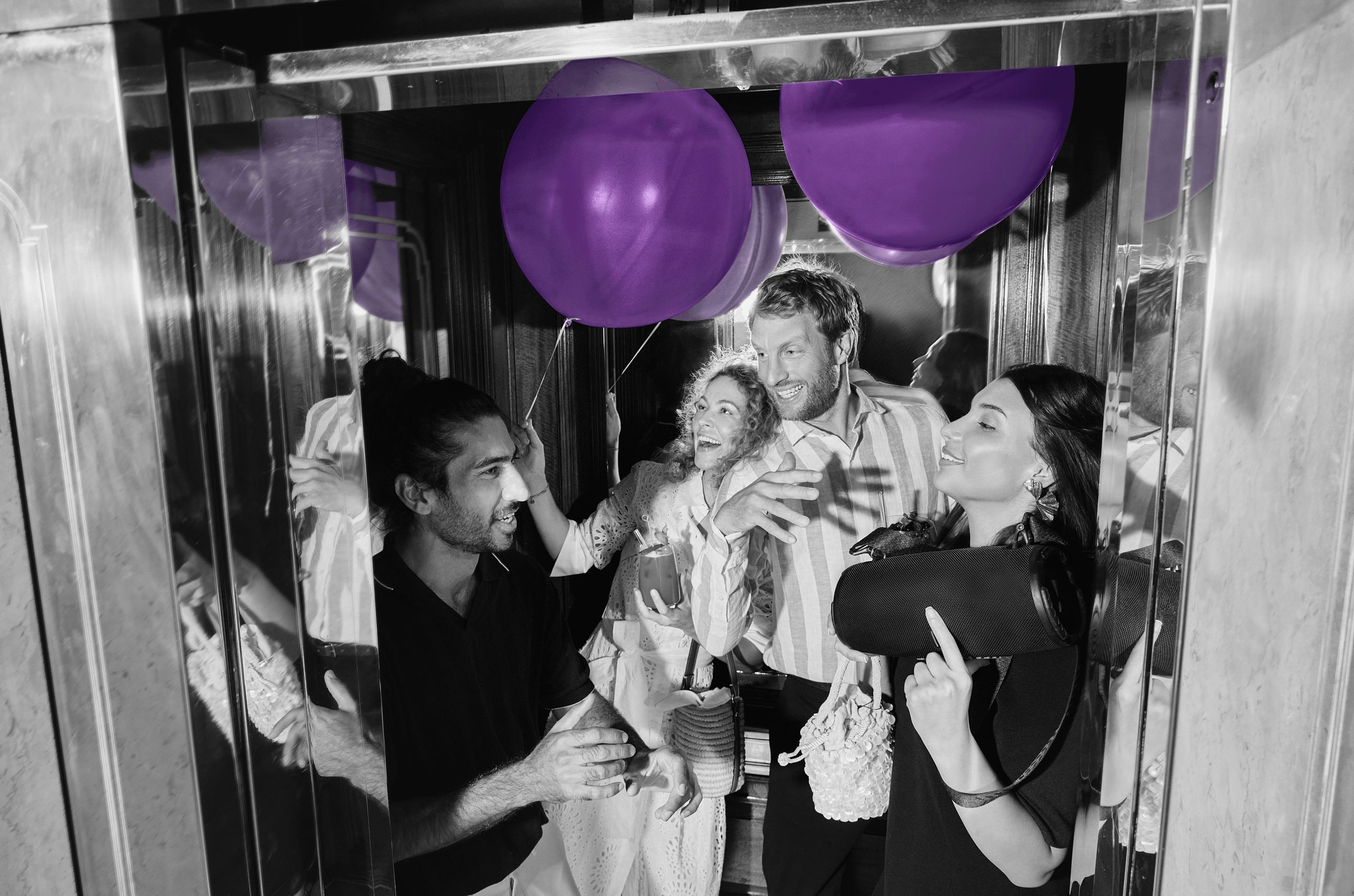 A group of people are inside an elevator, holding balloons and drinks, seemingly enjoying a social…