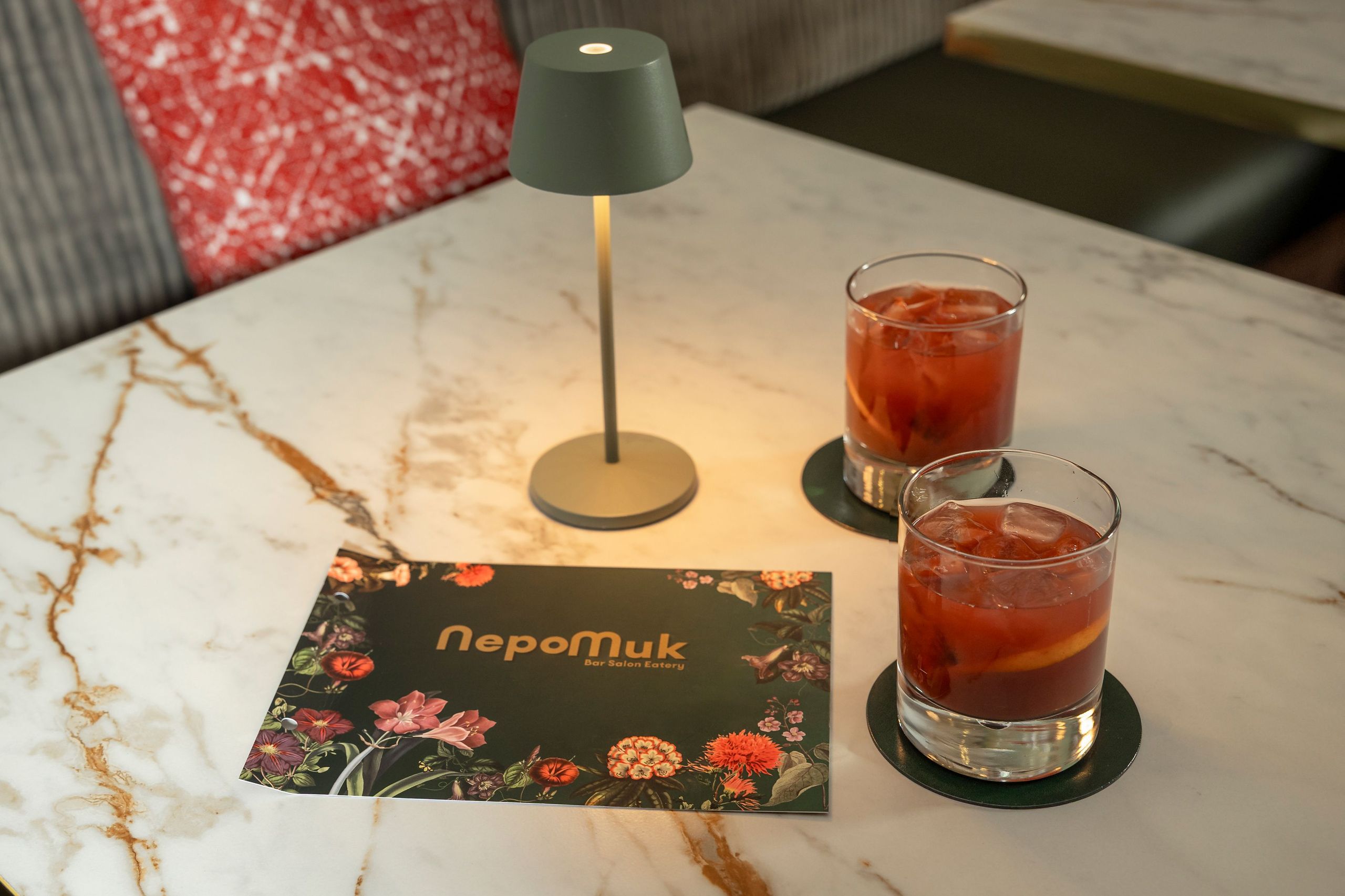 Un tavolo con una piccola lampada, un menu e due bicchieri di bevande rosse su sottobicchieri.