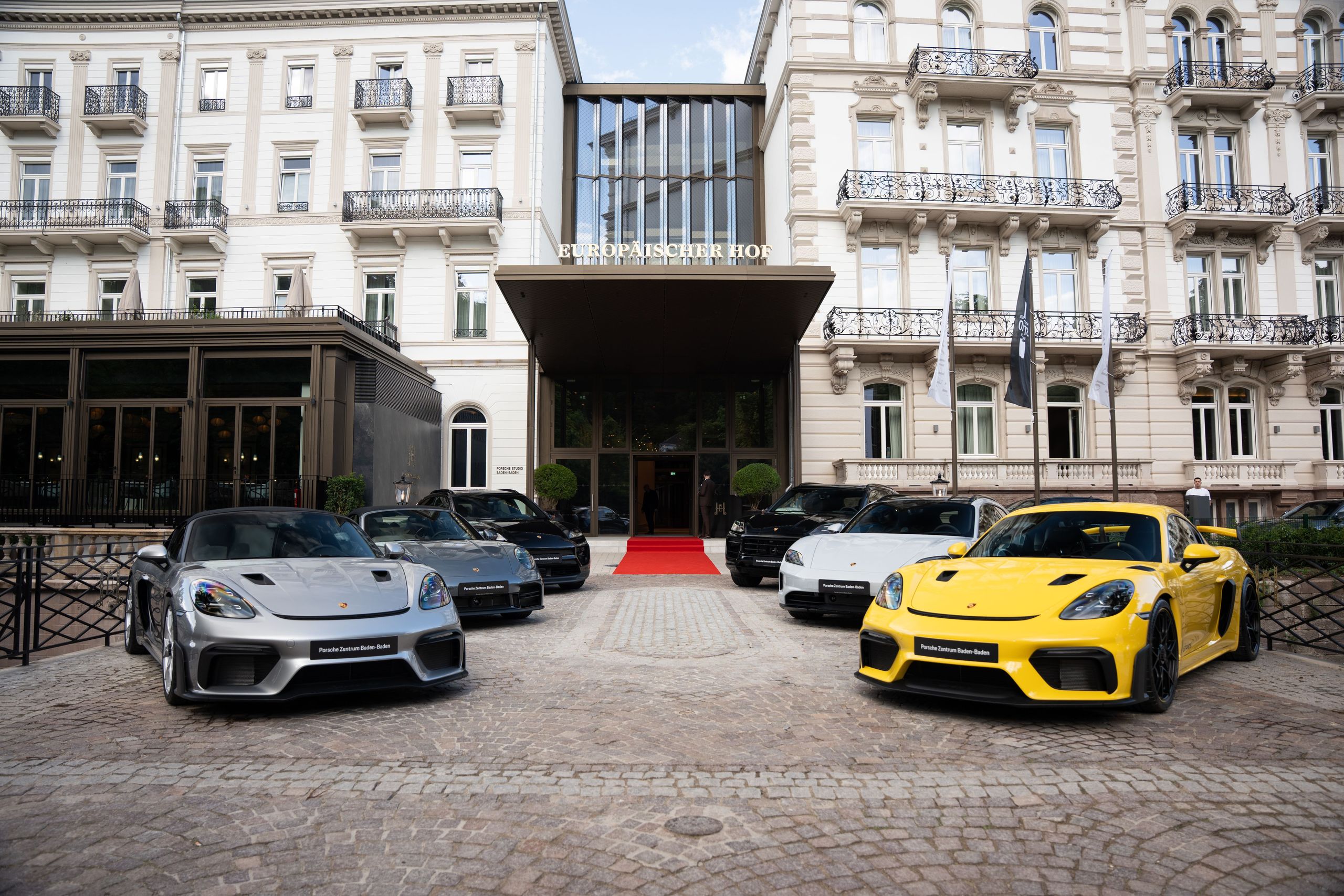 The photo shows a group of luxury sports cars parked in front of a grand hotel entrance.