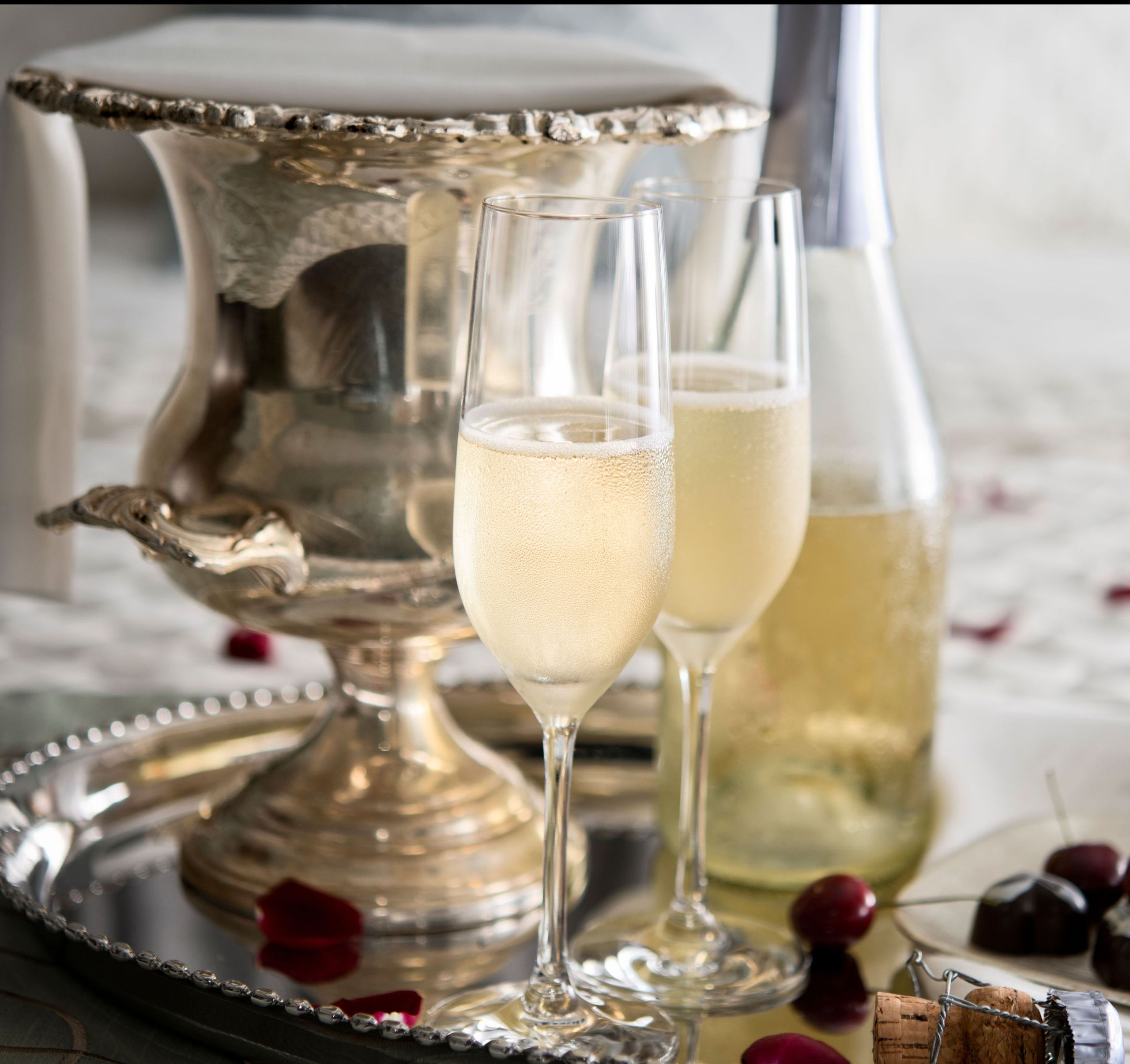 A silver ice bucket, a bottle of champagne, two filled champagne glasses, some cherries, and a…