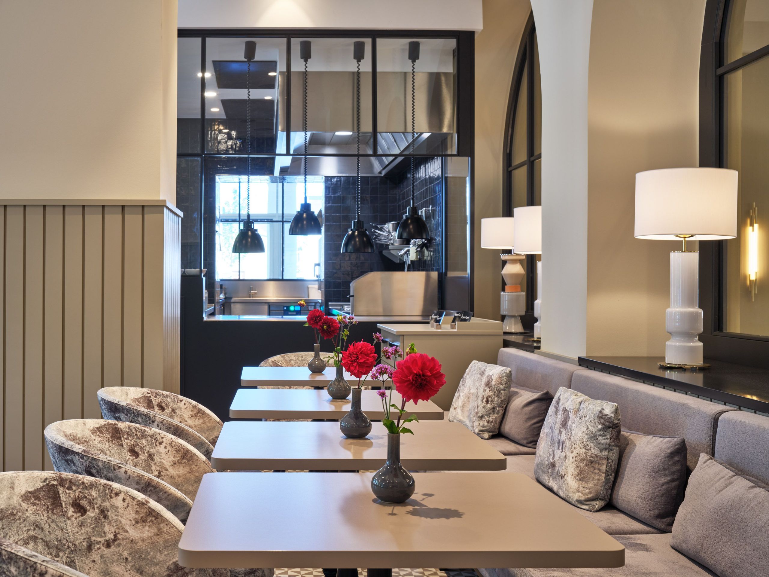 照片展示了一个现代餐厅区域，配有桌子、椅子和软垫长椅，装饰有装有红色花朵的花瓶和台灯，背景可见厨房区域。