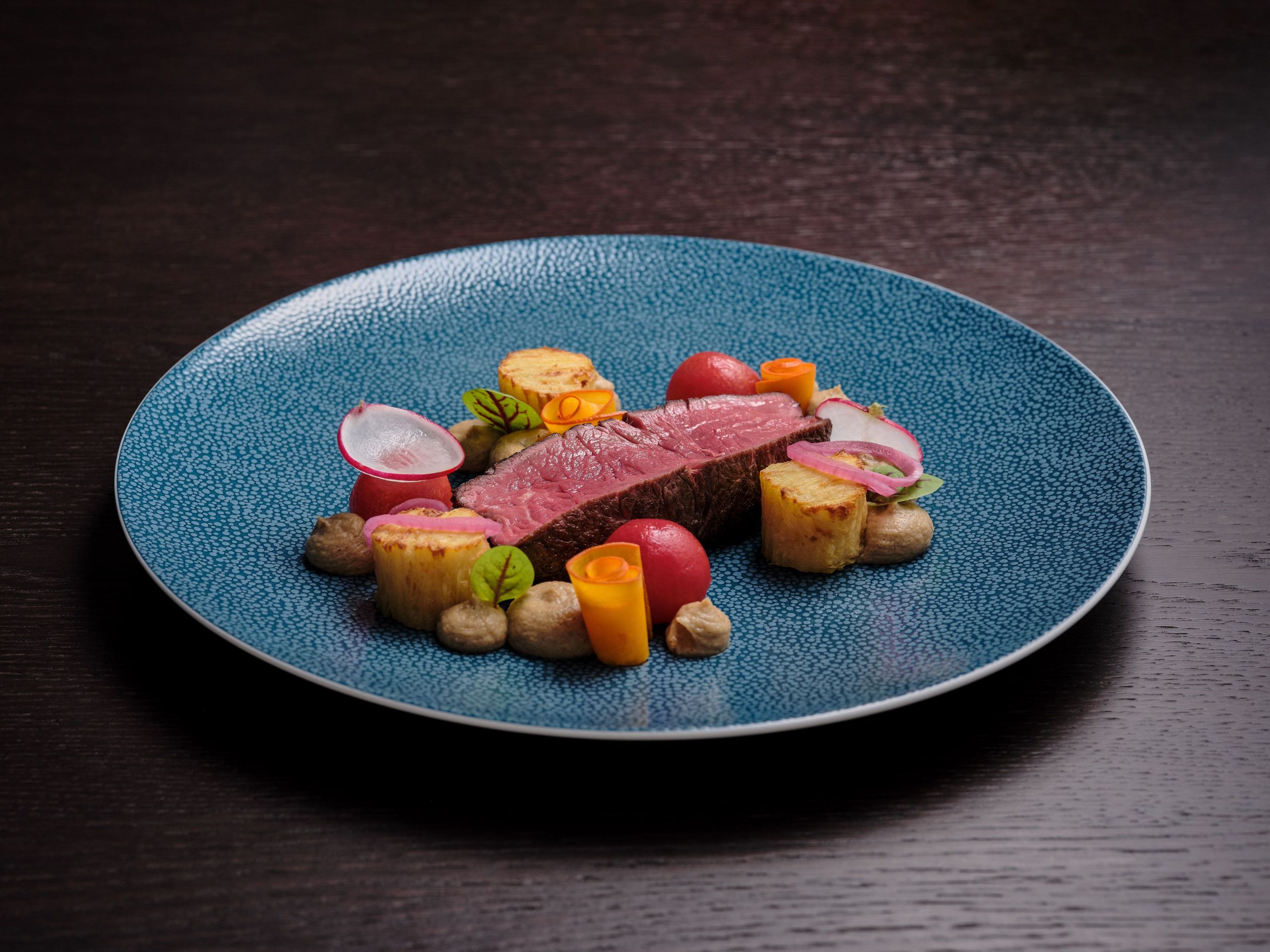 一份精致的菜肴，包含一块熟肉、各种蔬菜和装饰，摆放在蓝色的盘子上。