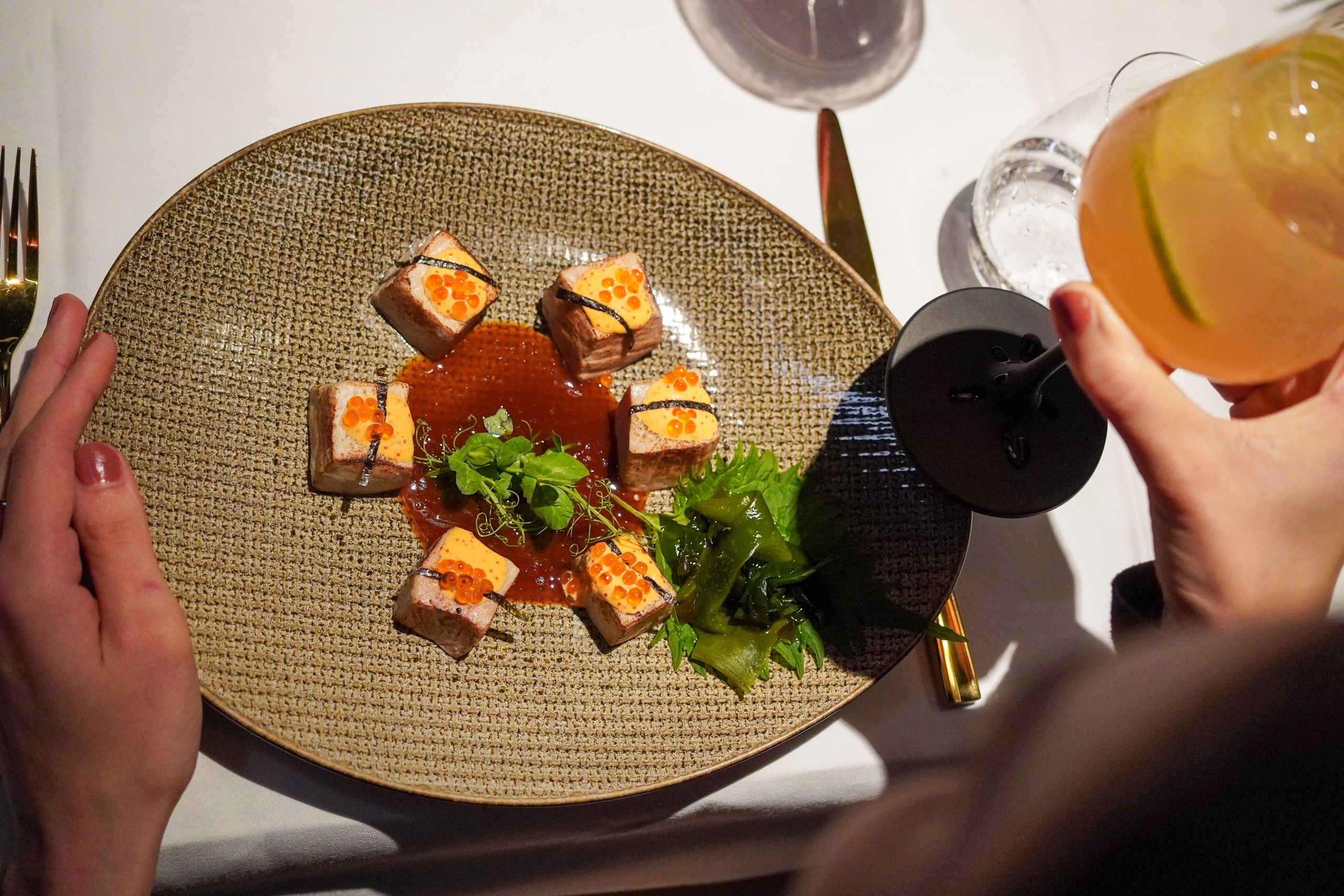Eine Person sitzt an einem Tisch mit einem Teller Gourmet-Essen und hält ein Glas mit einem Getränk.