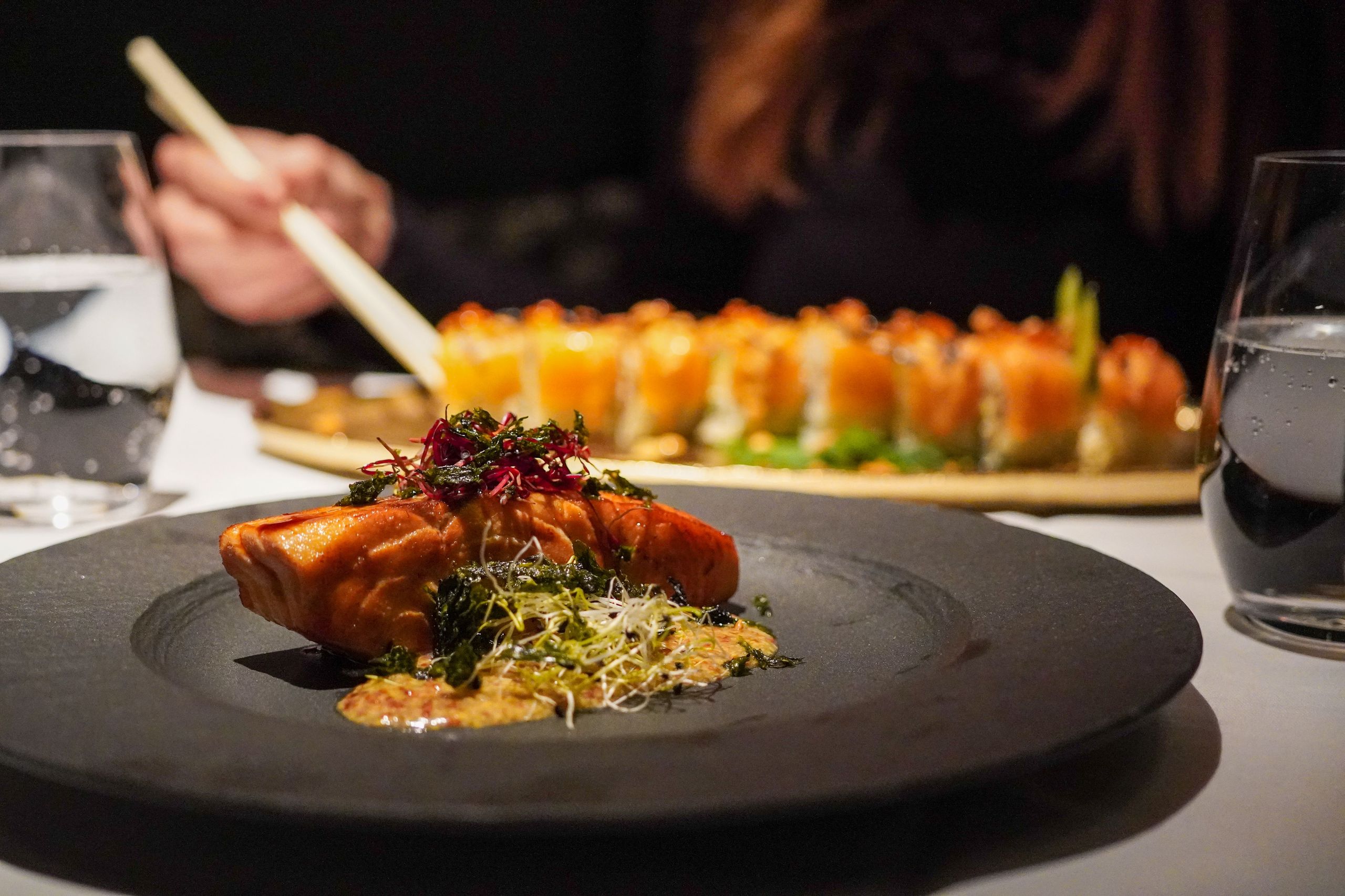 Na předním plánu talíř s kusem vařeného lososa a ozdobou, v pozadí rozmazané sushi a osoba…