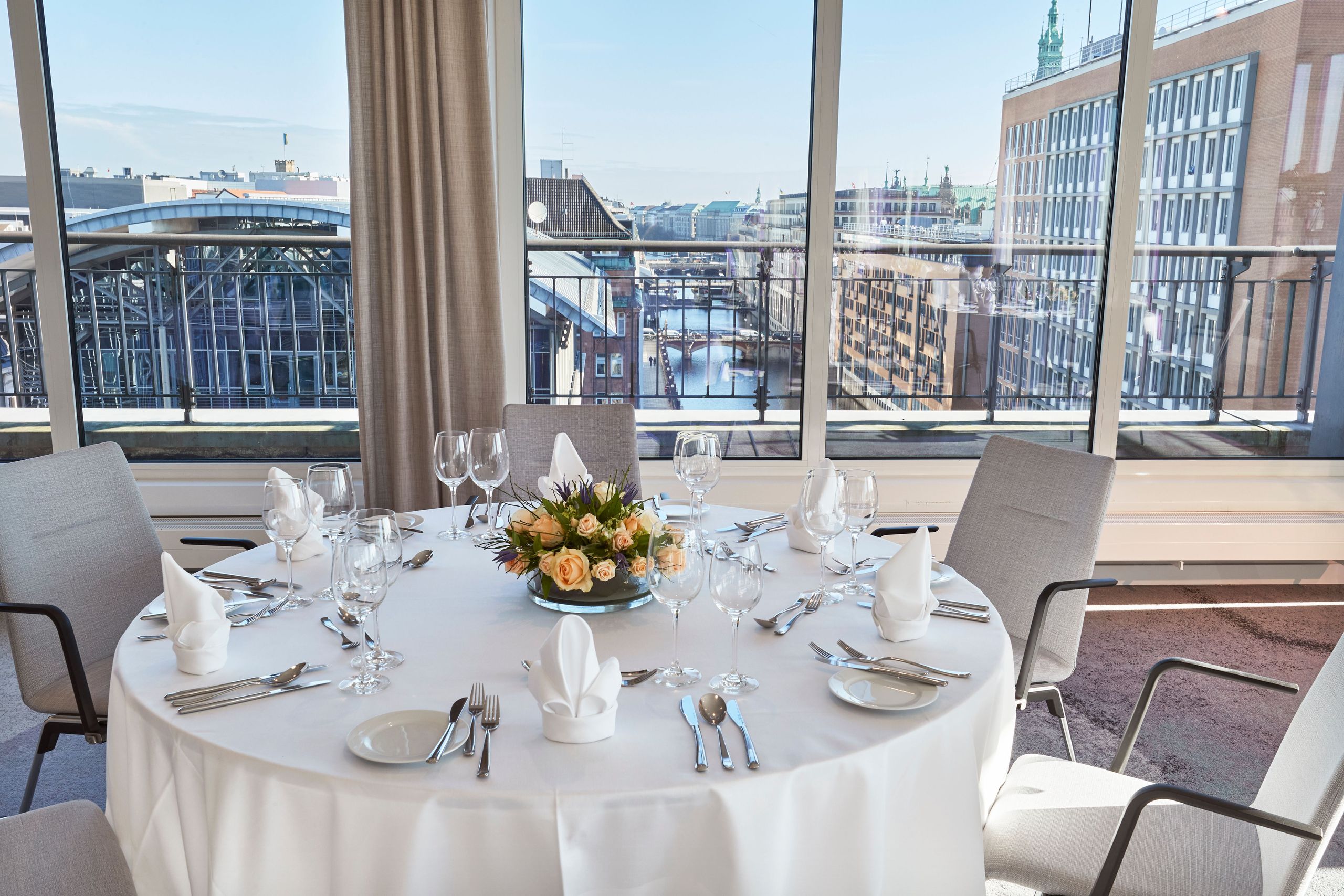 Ein runder Esstisch, gedeckt für eine Mahlzeit, mit Stühlen darum und einer Stadtsicht durch große…