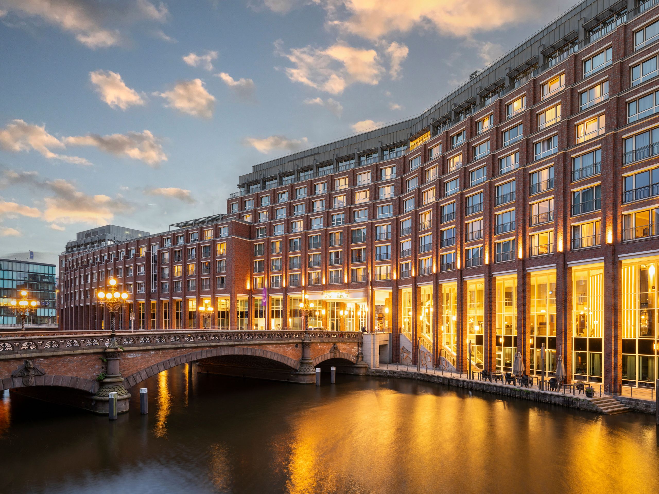 A cityscape featuring a large building illuminated at dusk next to a river with a bridge crossing…