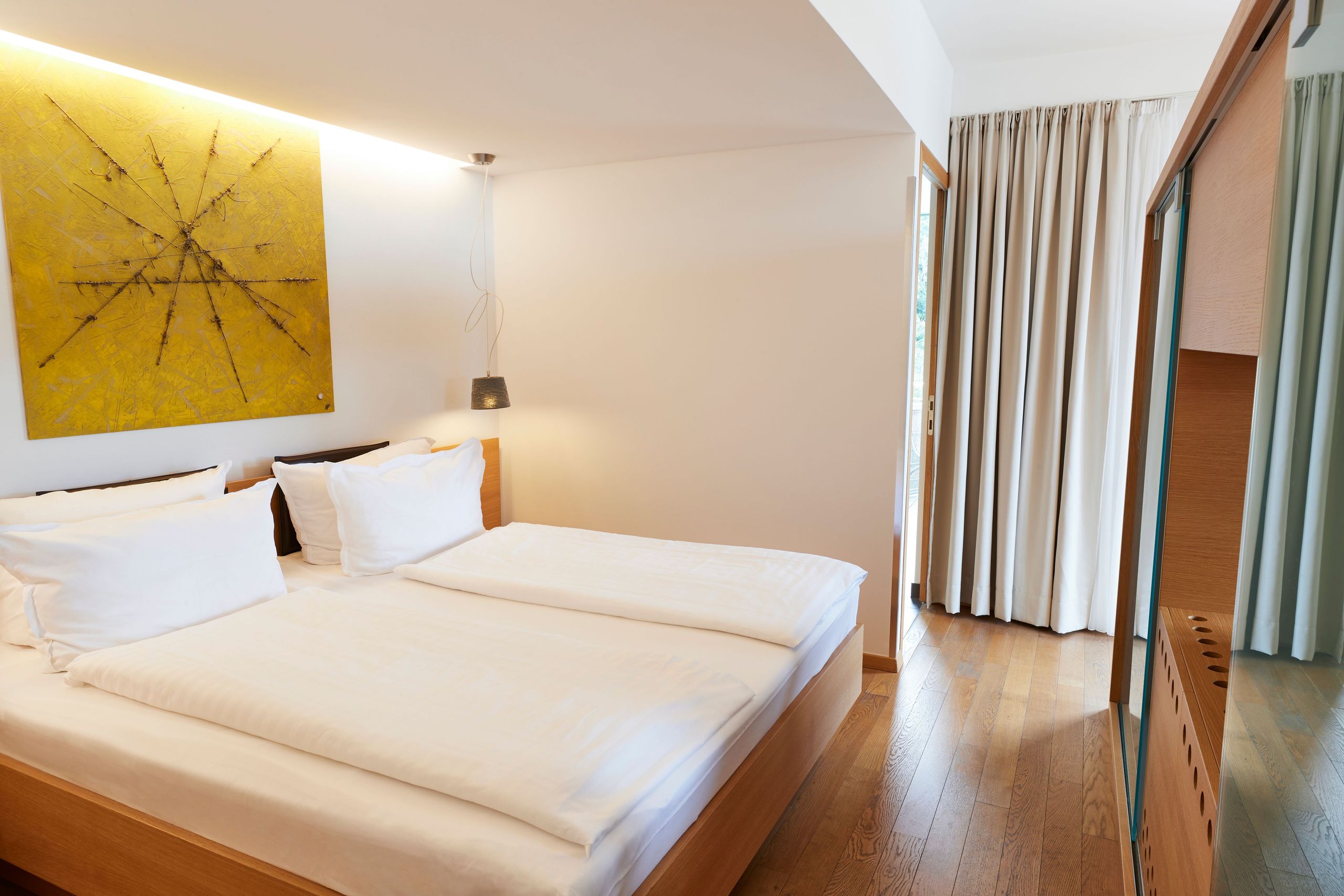 Un dormitorio con una cama doble, ropa de cama blanca, una obra de arte en la pared, una lámpara de pie, cortinas y un armario con espejo.