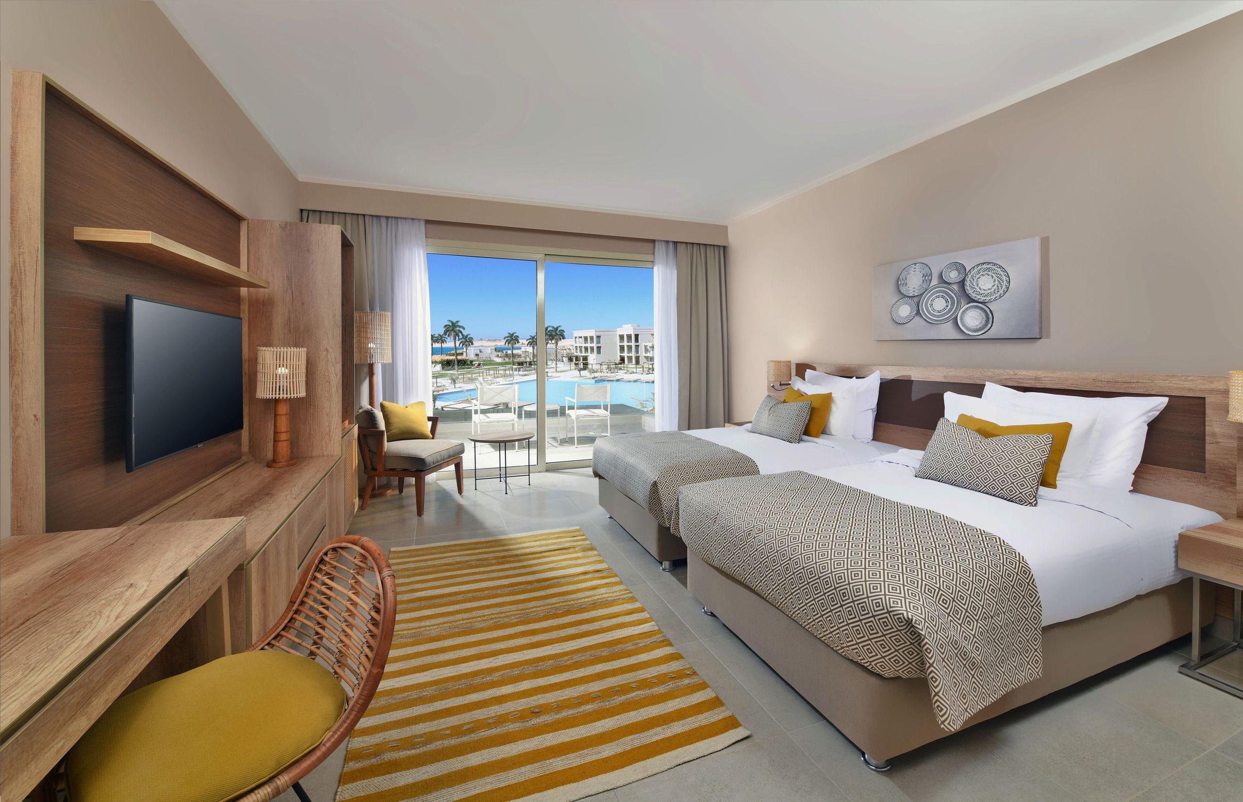 غرفة فندقية حديثة تحتوي على سريرين، تلفاز، كرسي، مكتب وشرفة تطل على مسبح ومبانٍ.