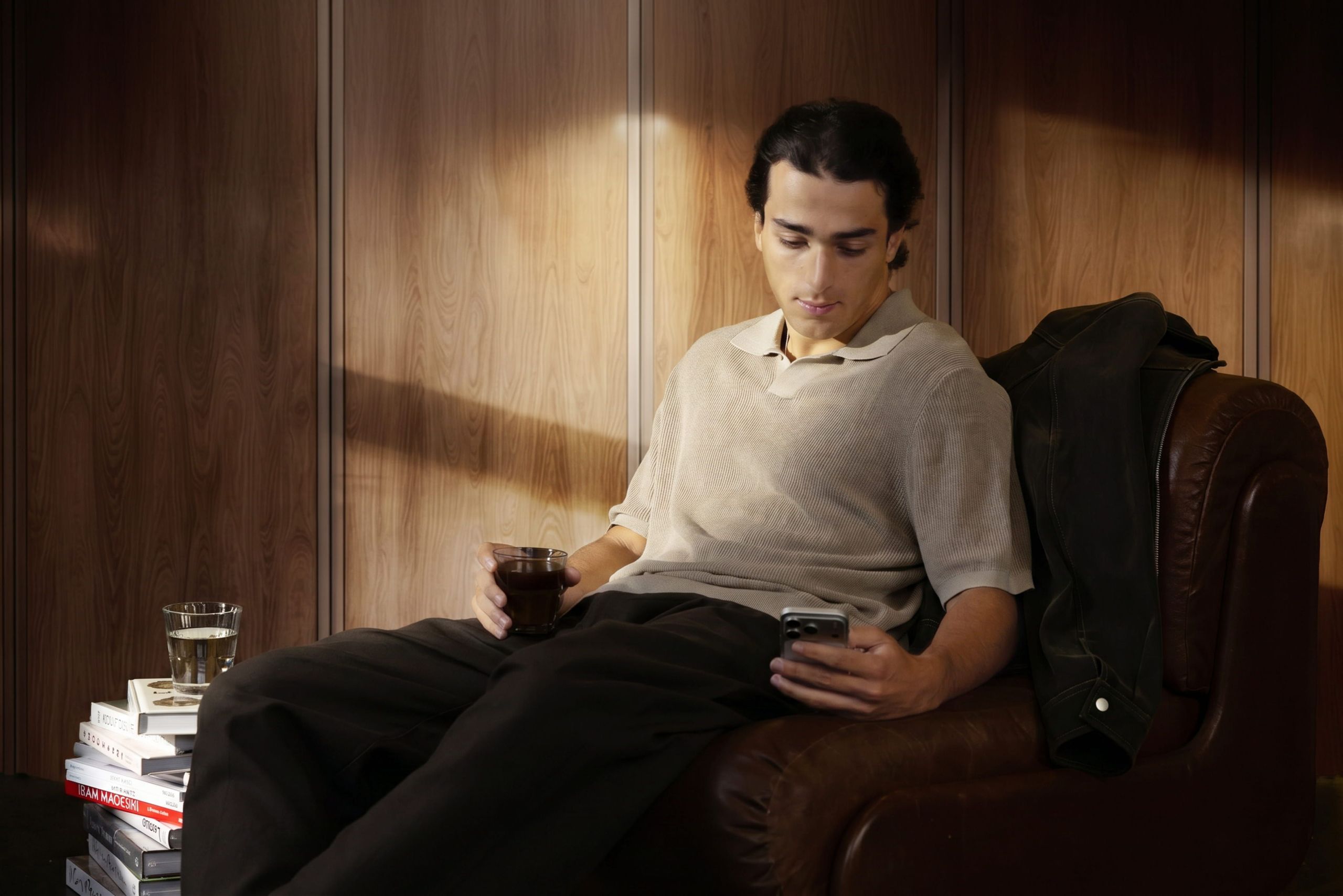 A man is sitting on a chair, holding a drink in one hand and looking at a phone in the other.