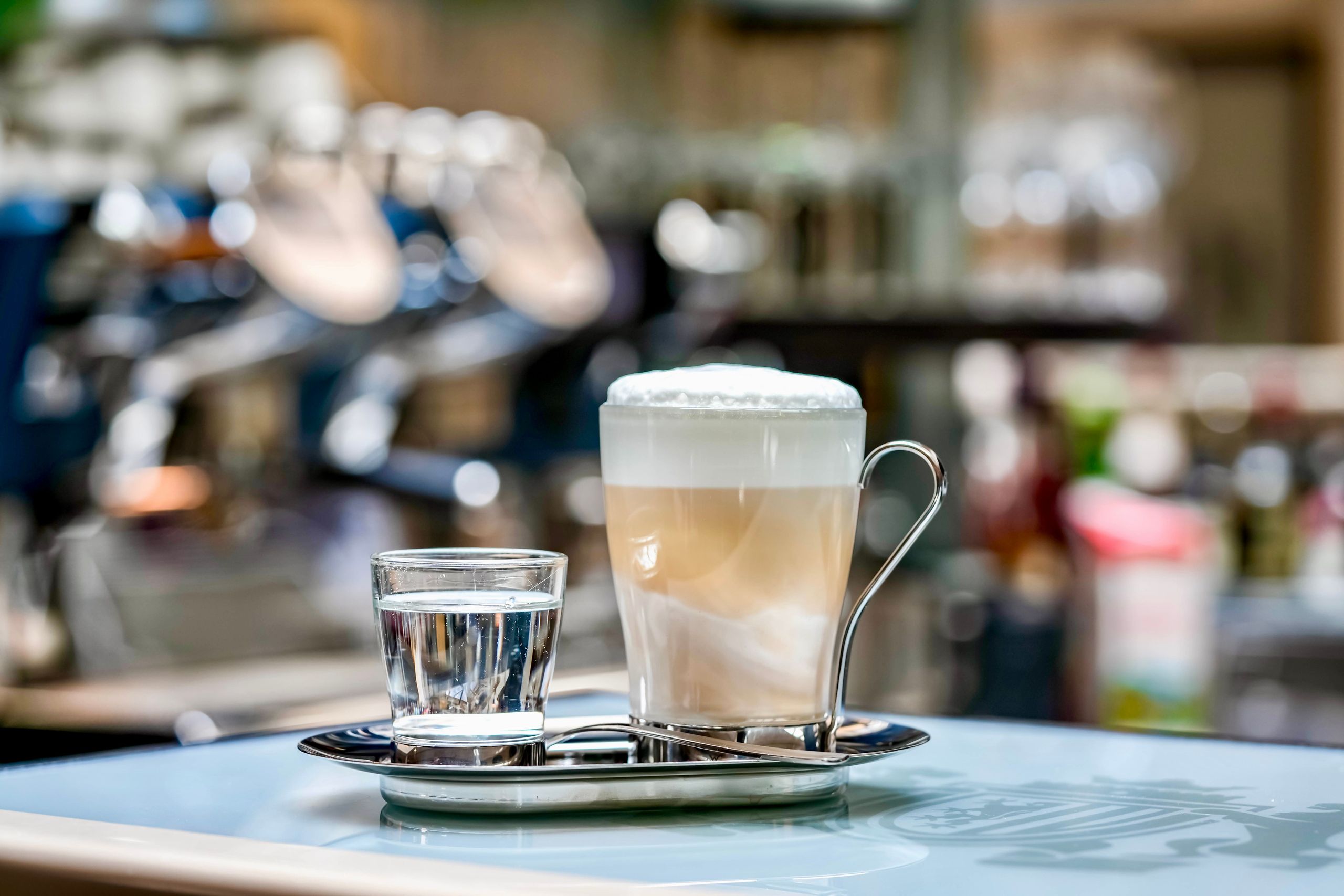 A glass of latte coffee with foam and a glass of water on a tray, placed on a table in a cafe…