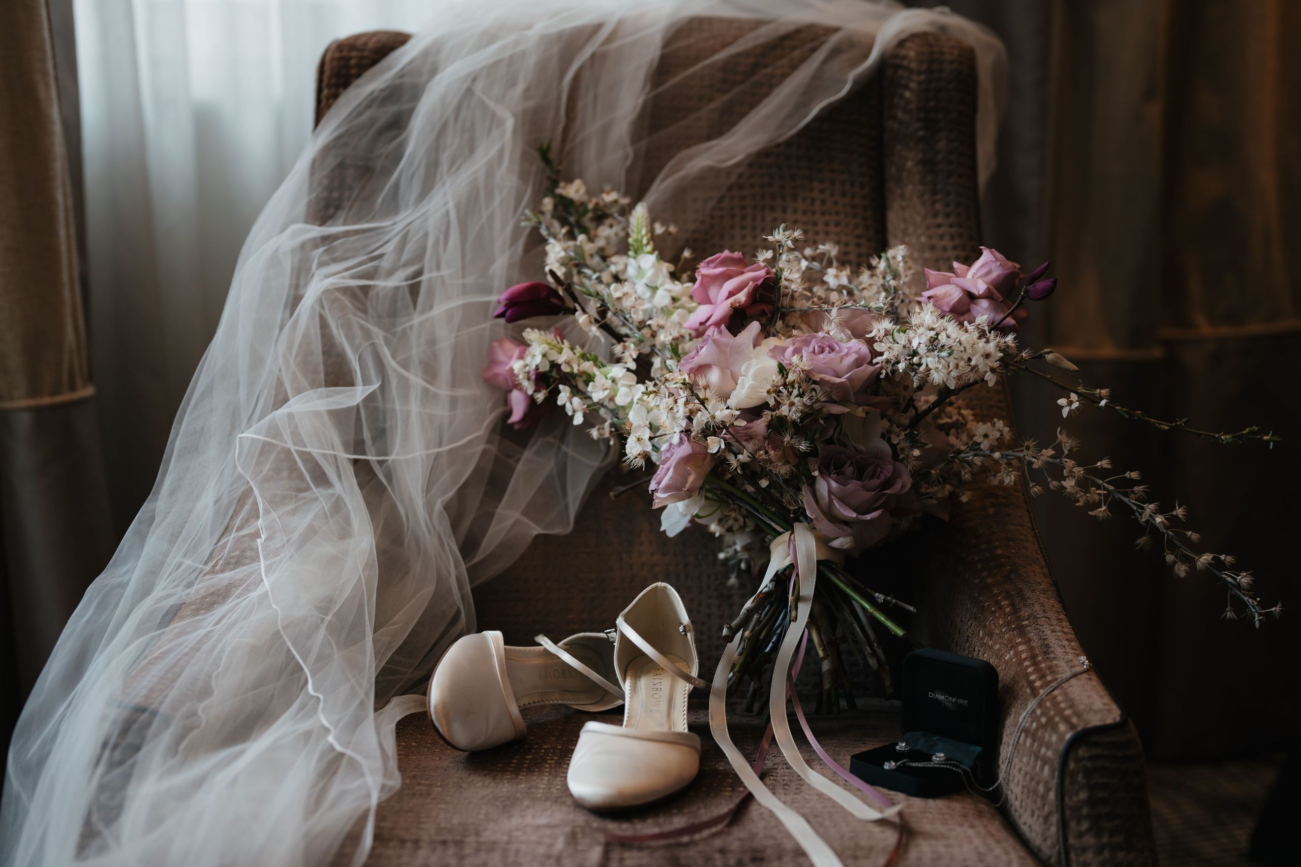 Das Foto zeigt einen Brautschleier, einen Blumenstrauß, ein Paar weiße Schuhe und eine Ringbox,…
