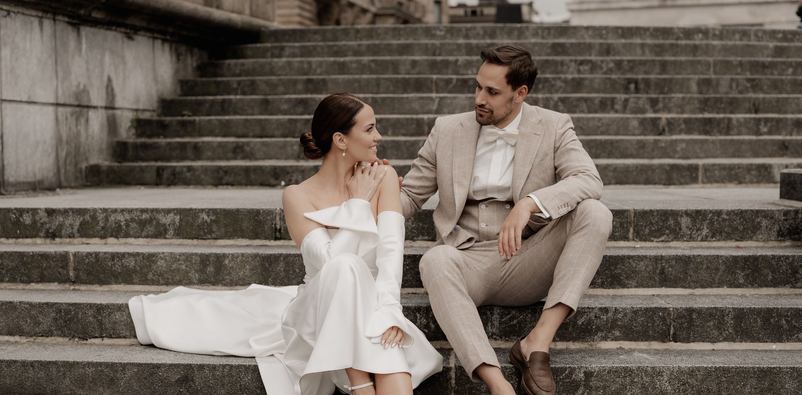 Пара в формальной свадебной одежде сидит на каменных ступенях на улице и смотрит друг на друга.