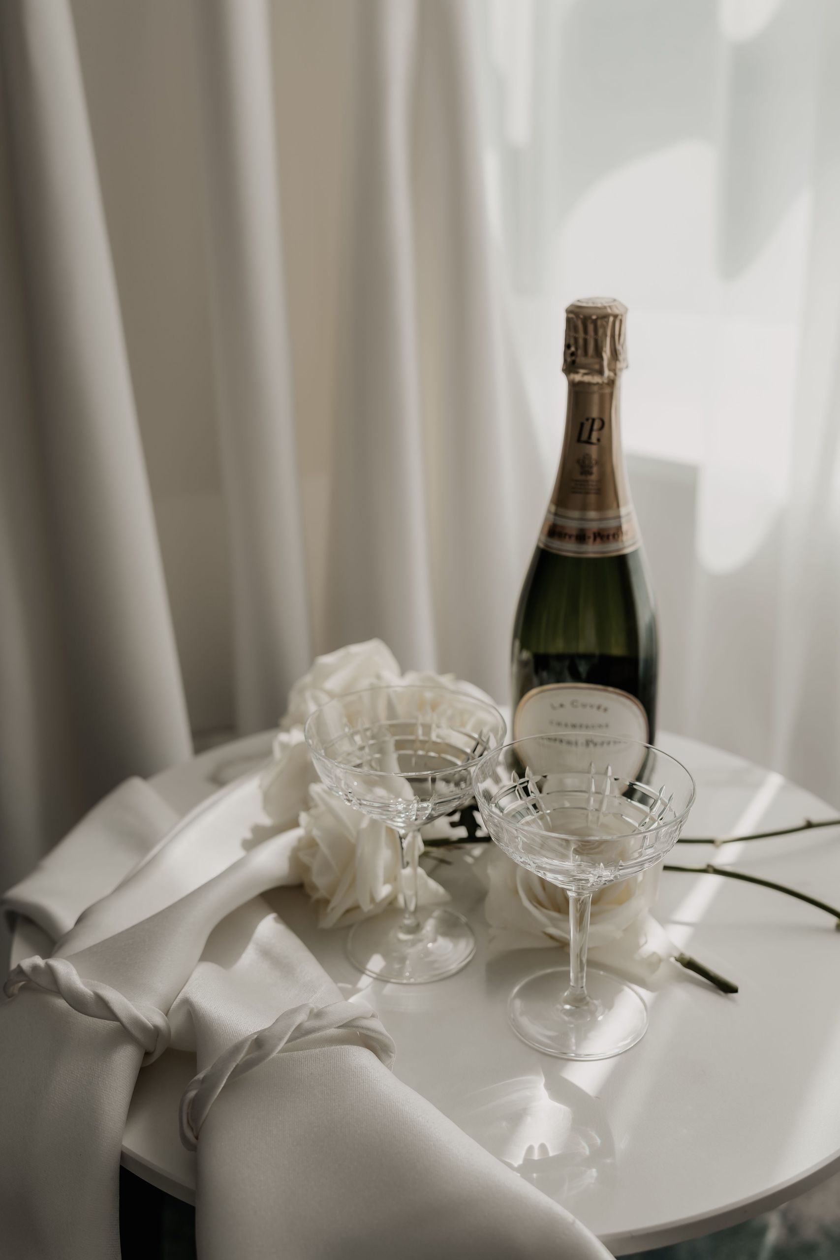 Бутылка шампанского, два хрустальных бокала и белые розы на столе с белой тканью.