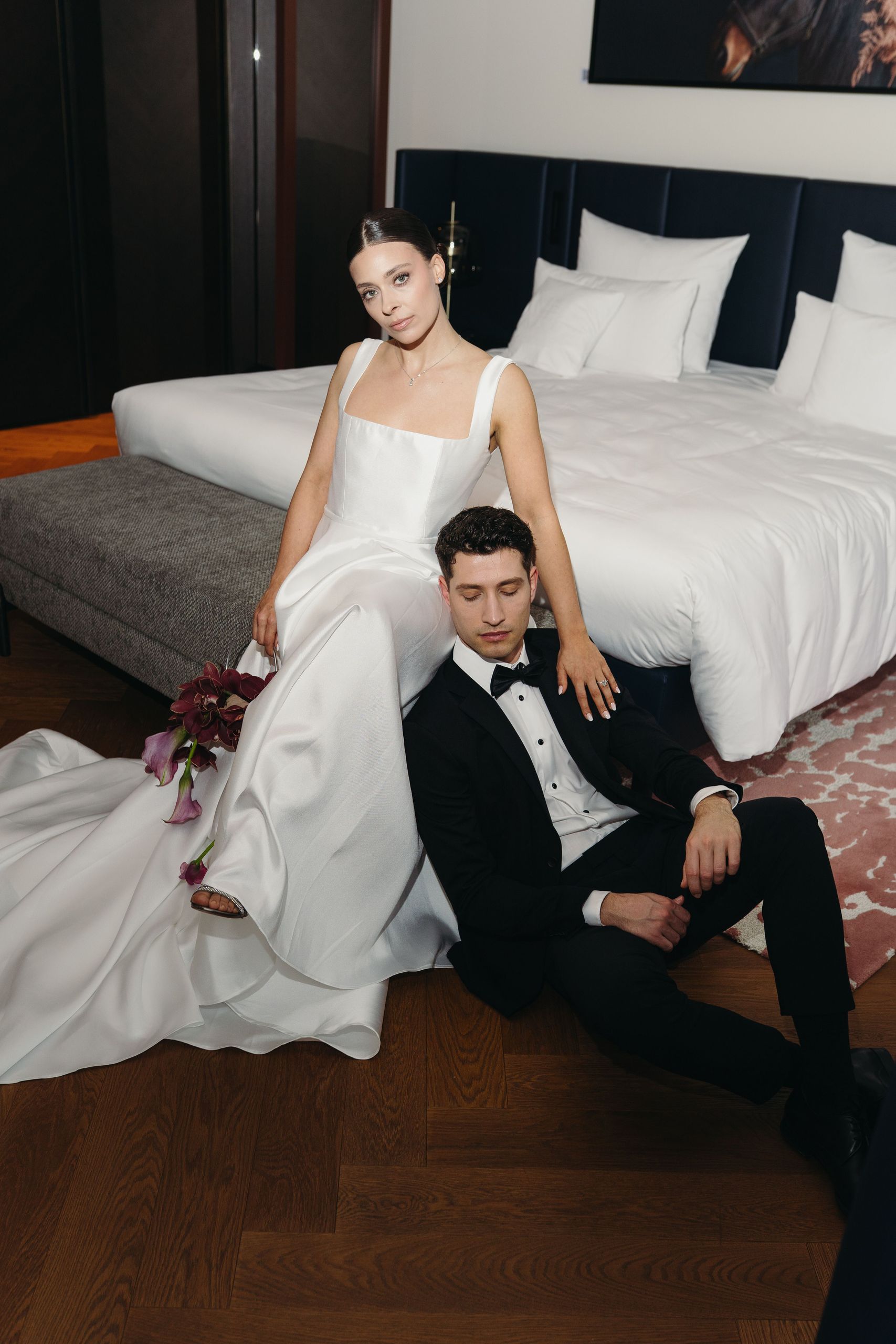 Жених и невеста в свадебной одежде позируют в спальне, невеста сидит на скамейке, а жених на полу.