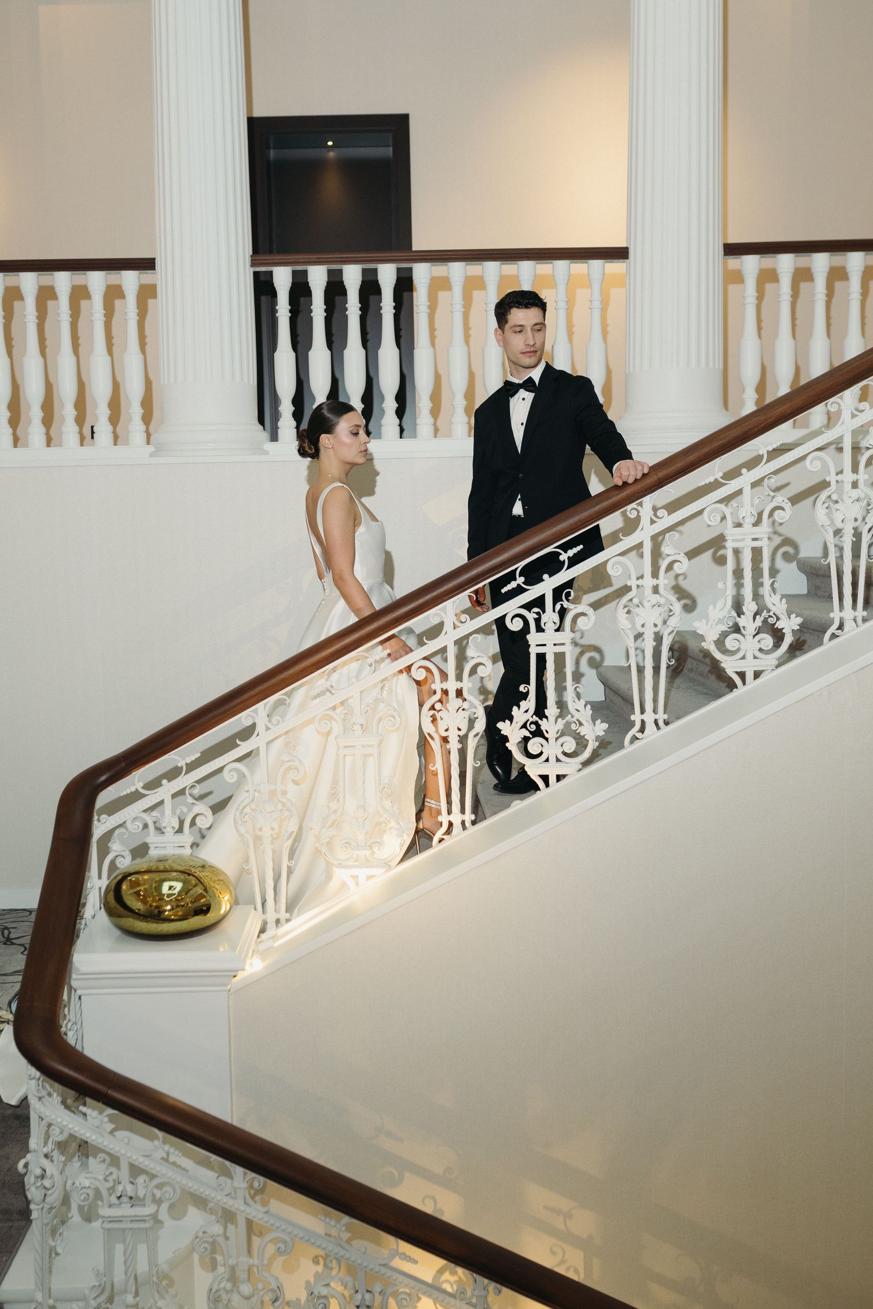 Пара в формальной одежде стоит на лестнице внутри здания с элегантными архитектурными элементами.
