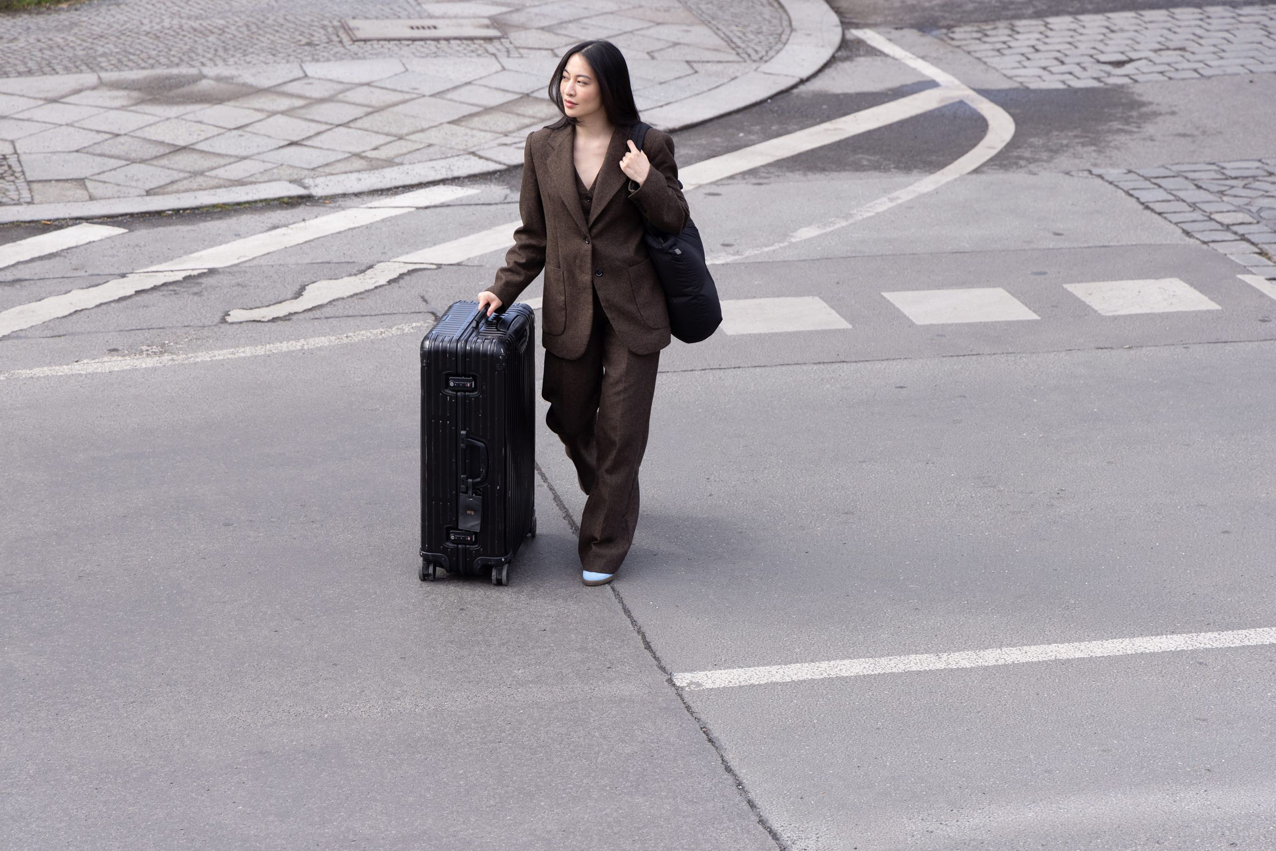 A woman in a suit is walking on a street while pulling a suitcase.