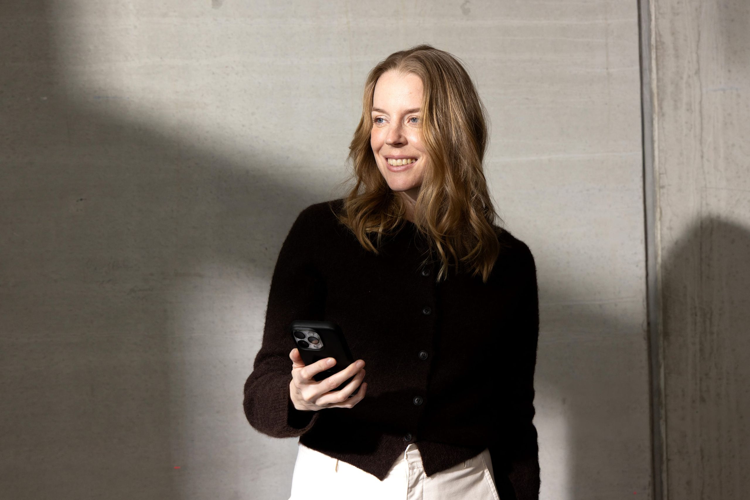 A woman holding a smartphone and smiling, standing against a plain background.