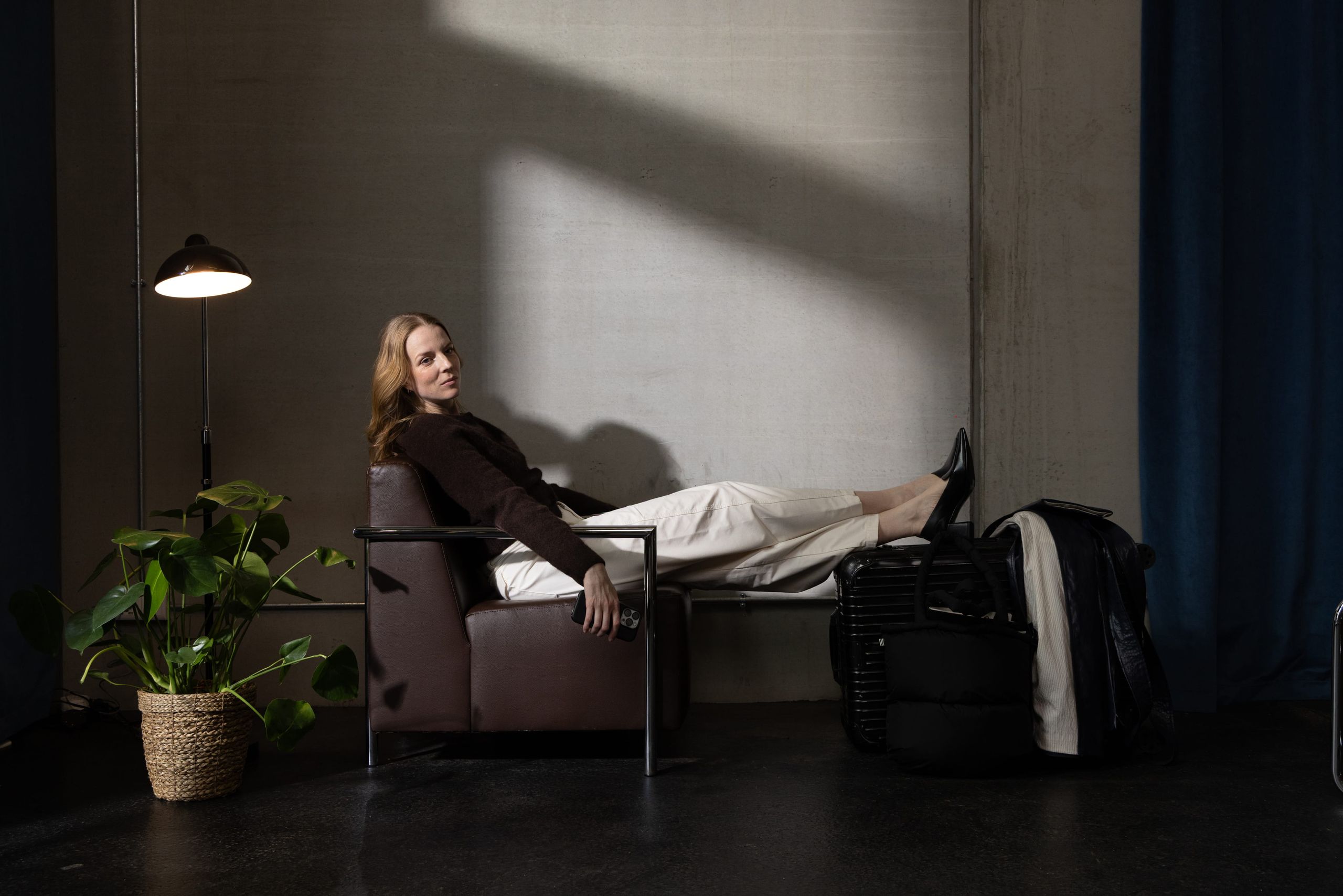 A woman is sitting relaxed on a chair with her legs stretched out on a suitcase in a room with dim…