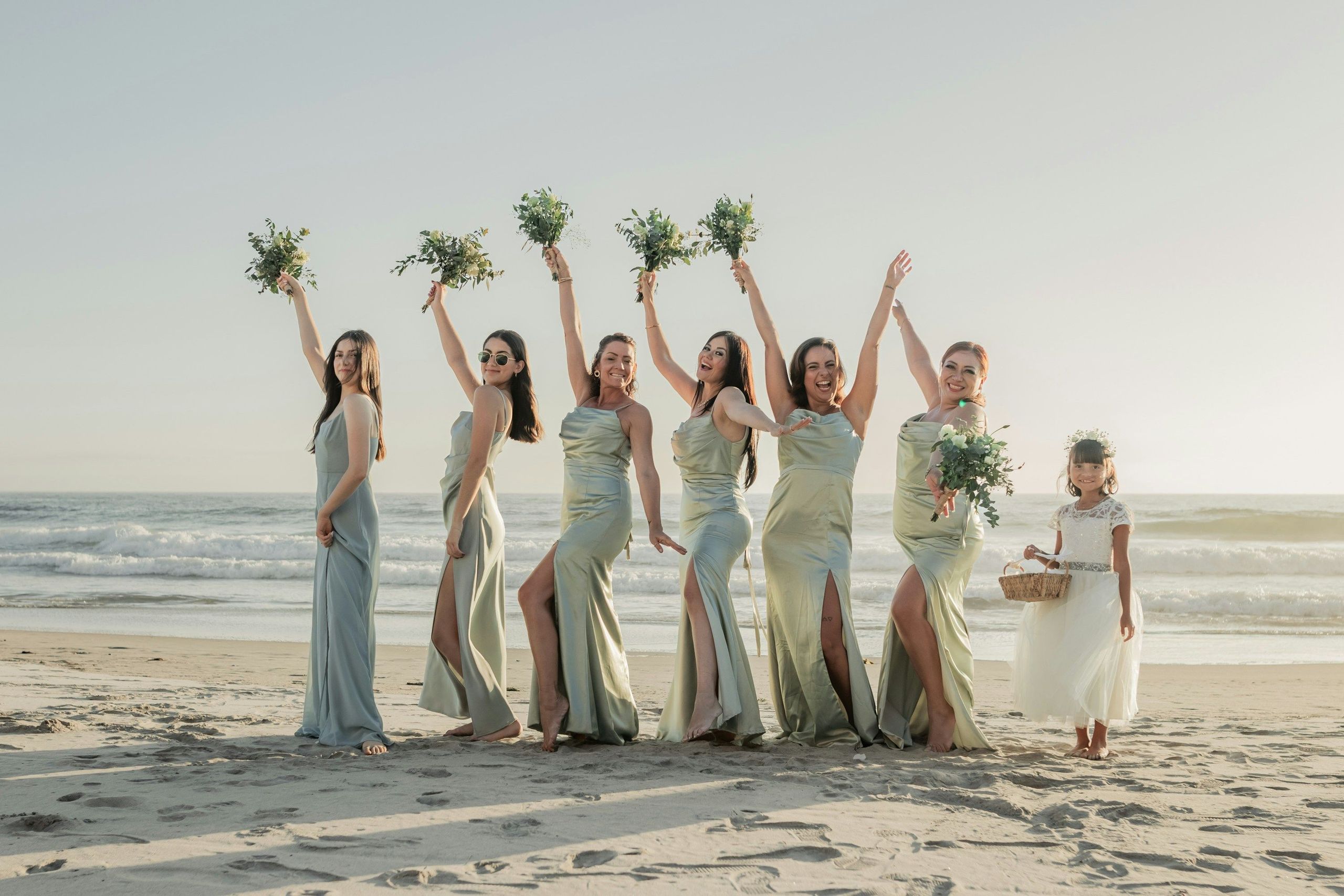 مجموعة من النساء وفتاة صغيرة يرتدين ملابس رسمية يقفن على الشاطئ، والنساء يرفعن أذرعهن ويمسكن باقات…