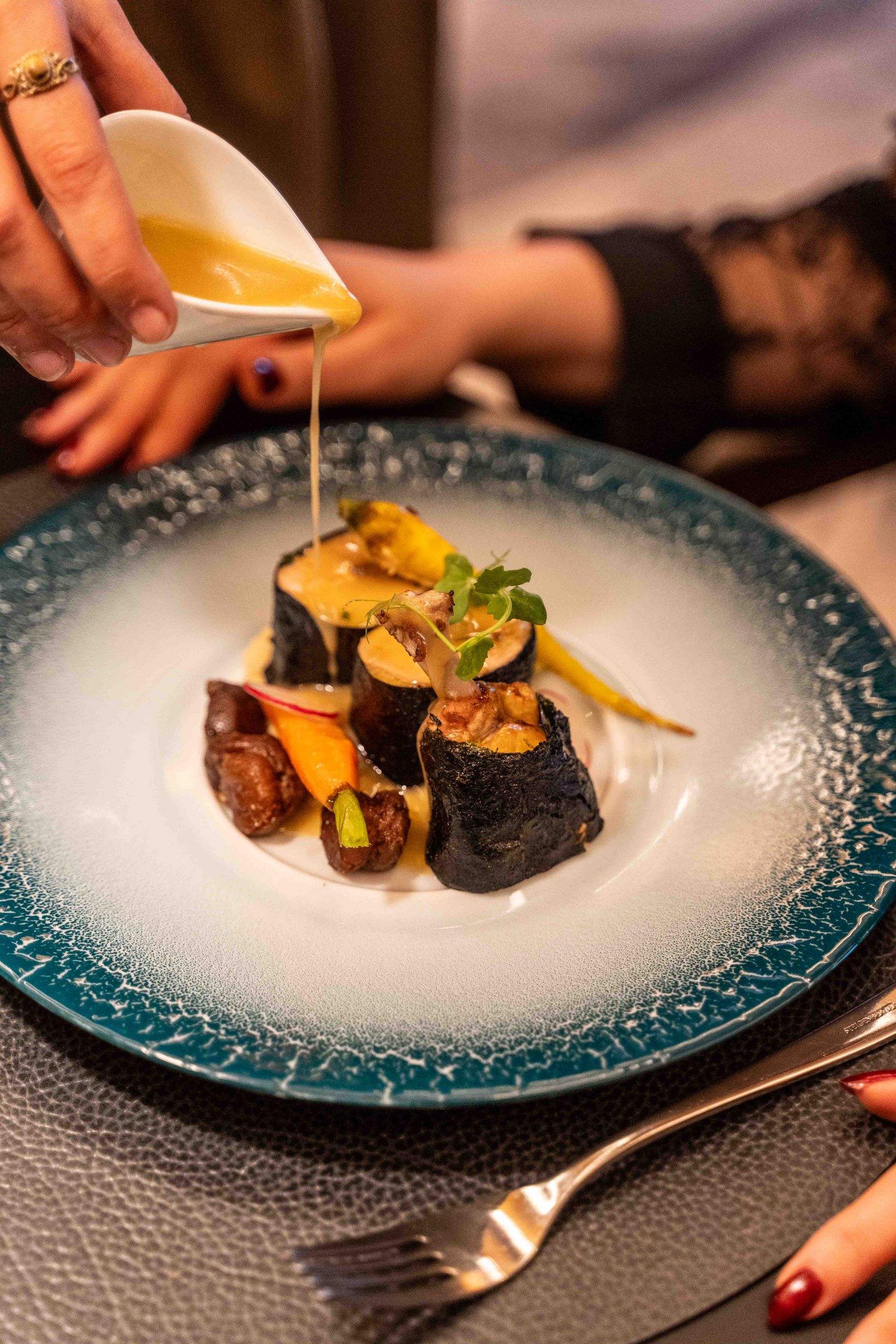 Une personne verse une sauce sur un plat gastronomique sur une assiette décorative.